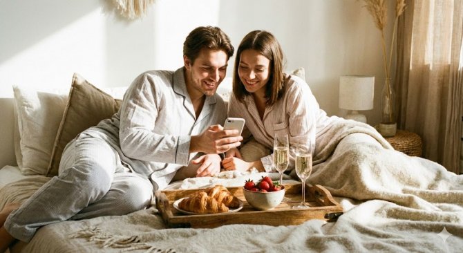 Pohodov&eacute; r&aacute;no v posteli: P&aacute;r společně sleduje mobil a vychutn&aacute;v&aacute; si čerstv&eacute; croissanty a jahody doručen&eacute; službou Rohl&iacute;k.cz. Ilustrace využit&iacute; slevov&eacute;ho k&oacute;du na prvn&iacute; n&aacute;kup na Rohl&iacute;k.cz pro dokonalou valent&yacute;nskou sn&iacute;dani do postele.