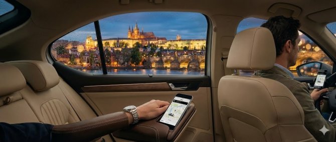 Pohled ze zadn&iacute;ho sedadla luxusn&iacute;ho vozu během j&iacute;zdy s Uberem v Praze. Pasaž&eacute;r sleduje aplikaci na telefonu, zat&iacute;mco řidič ř&iacute;d&iacute;. Z de&scaron;tiv&eacute;ho okna je večern&iacute; v&yacute;hled na osvětlen&yacute; Pražsk&yacute; hrad a Karlův most