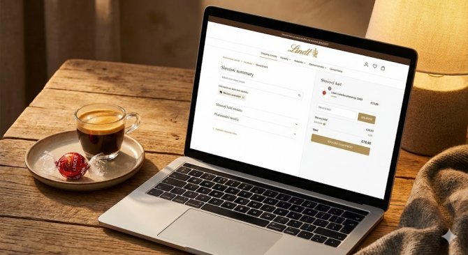 Detailní záběr na obrazovku notebooku s otevřeným košíkem e-shopu Lindt.cz a polem pro zadání slevového kódu, vedle šálku kávy a rozbalené pralinky Lindor.